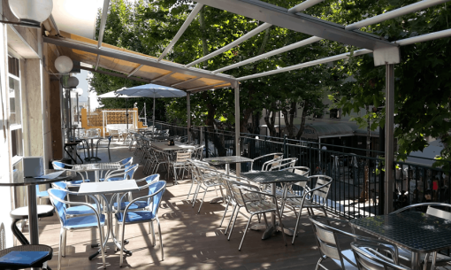 Terrazza caffè con sedie e tavoli in metallo, ombreggiata da alberi.